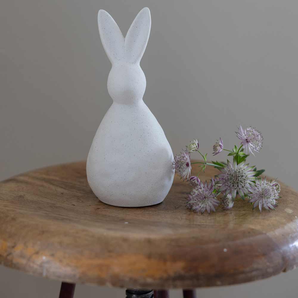 Storefactory - Alma Hase Figur neben lila Blumen auf einem Holzhocker.