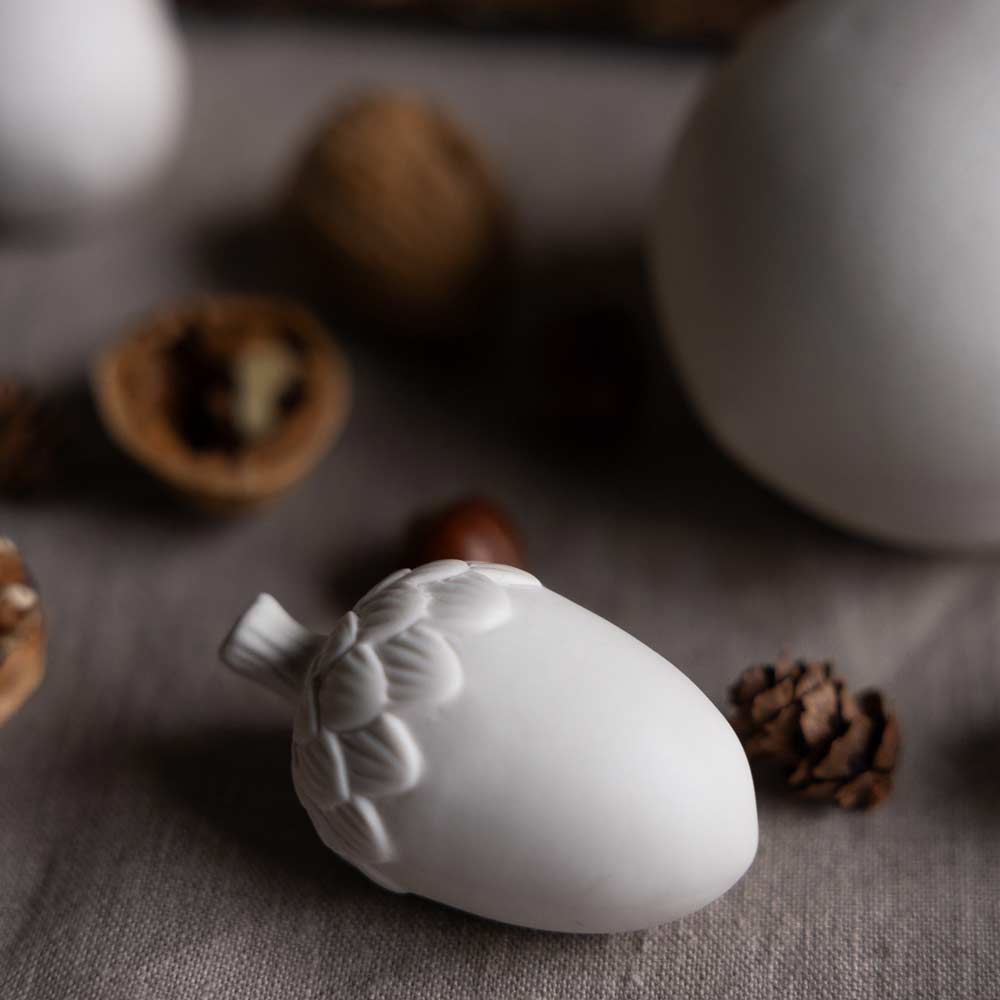 Die Storefactory – Eichel Keramik, ein weißes Eichel-Ornament aus Keramik, wird auf einer Stoffoberfläche inmitten natürlicher Elemente, darunter Walnüsse und Tannenzapfen, im Hintergrund präsentiert.