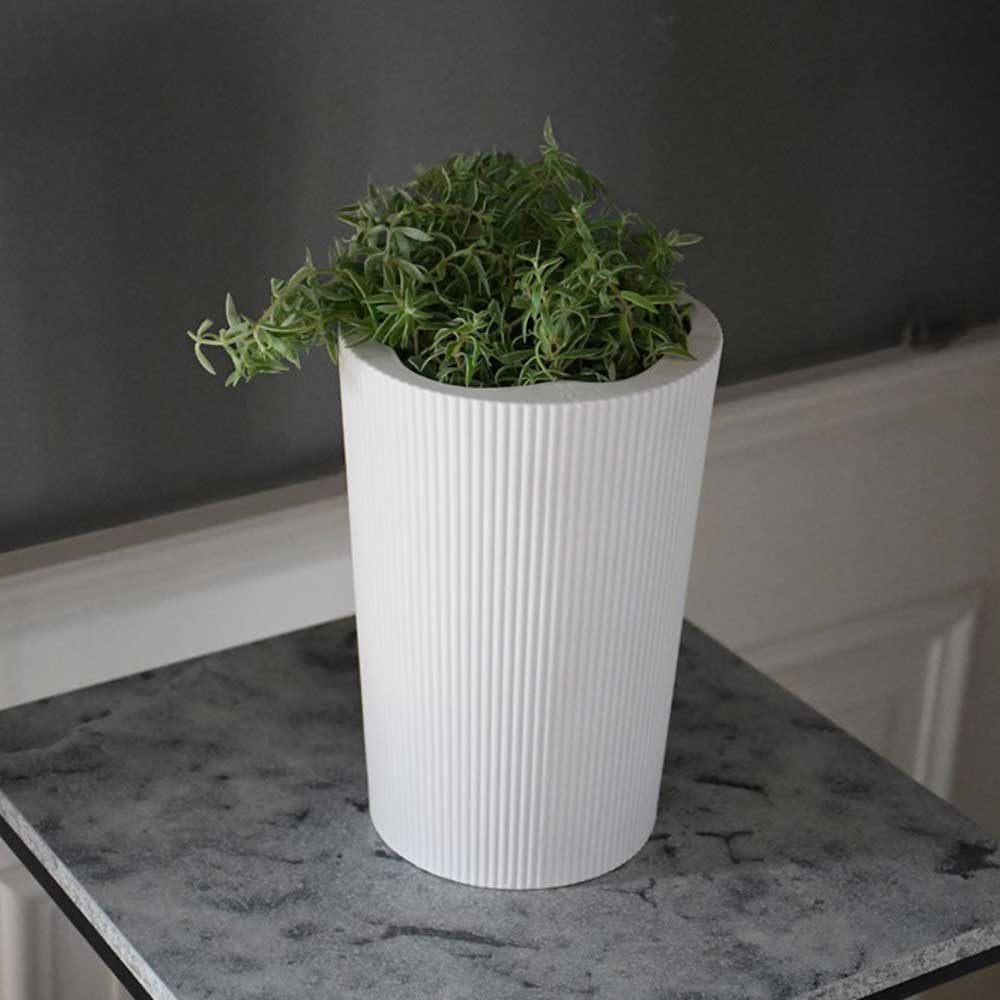 Die Eksberg Vase von Storefactory, ein weißer, gerippter, zylindrischer Topf, hält eine grüne Pflanze auf einer gesprenkelten grauen Oberfläche vor einer dunklen Wand.