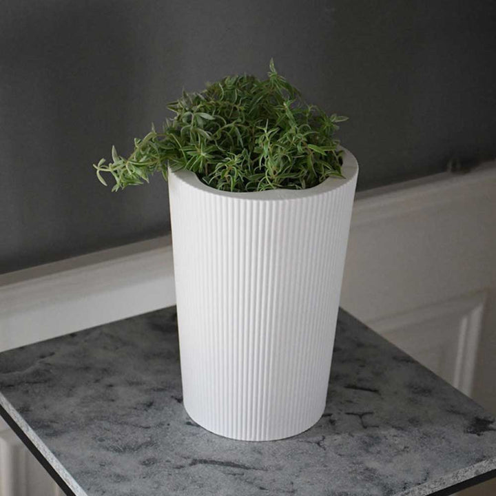 Die Eksberg Vase von Storefactory, ein weißer, gerippter, zylindrischer Topf, hält eine grüne Pflanze auf einer gesprenkelten grauen Oberfläche vor einer dunklen Wand.