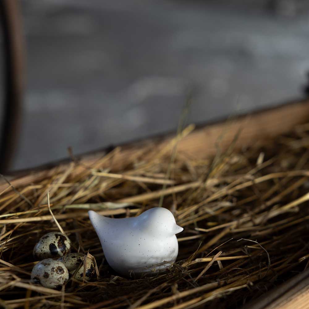 In einer schwach beleuchteten Umgebung zeigt die Storefactory – Ellen Huhn einen kleinen Keramikvogel mit drei gesprenkelten Eiern, der auf Stroh ruht.