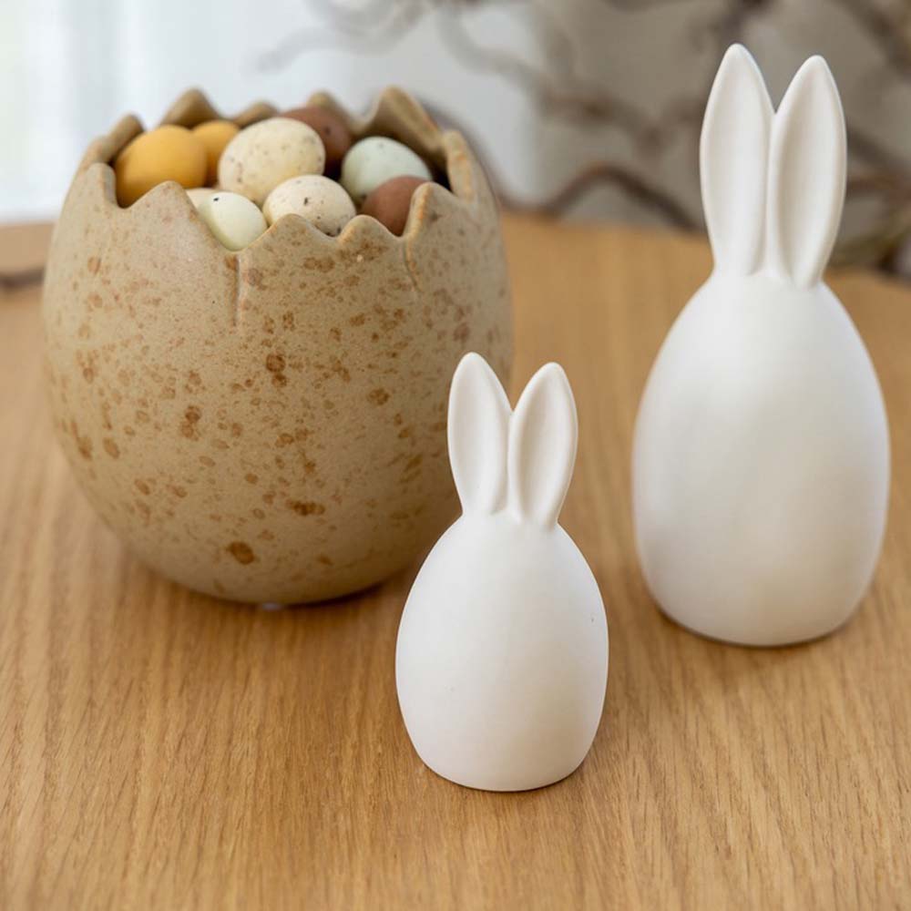Eierförmige Häschen aus Keramik von Hase Emma auf einer Holzoberfläche neben einer gesprenkelten Schüssel gefüllt mit kleinen Eiern.