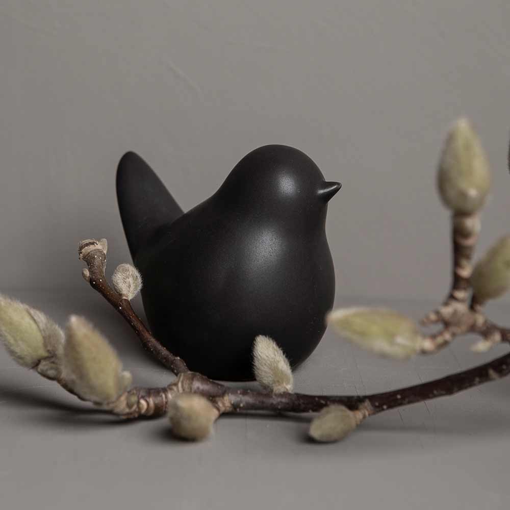 Eine Vogelfigur von Storefactory - Herman Vogel, die auf einem Ast mit knospenden Weidenkätzchen vor einem grauen Hintergrund thront.