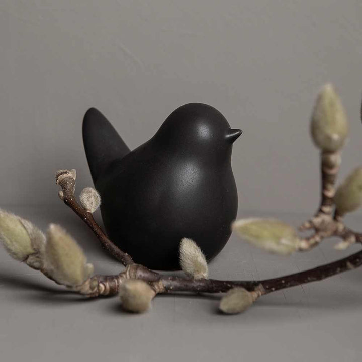 Eine Vogelfigur von Storefactory - Herman Vogel, die auf einem Ast mit knospenden Weidenkätzchen vor einem grauen Hintergrund thront.