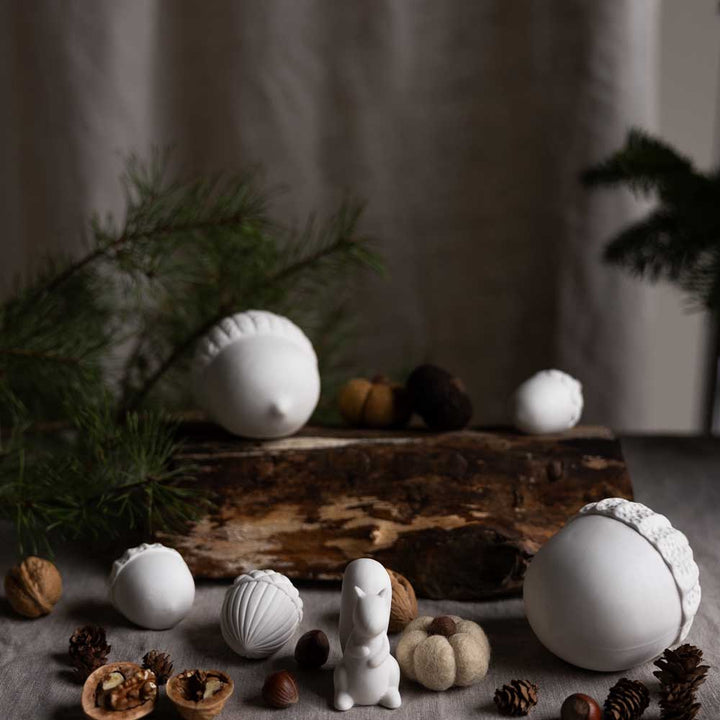 Storefactory – Keramik-Eicheln und eine kleine Hasenfigur von Holeby Eichel auf einem Tisch mit Tannenzweigen und verschiedenen Nüssen, vor einem neutralen Hintergrund.