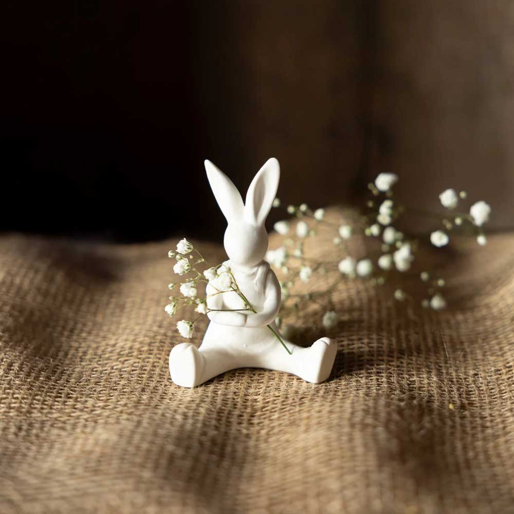 Die Storefactory – Lasse Hase ist eine weiße Hasenfigur aus Keramik, die auf strukturiertem Sackleinen sitzt und kleine Blumen hält.
