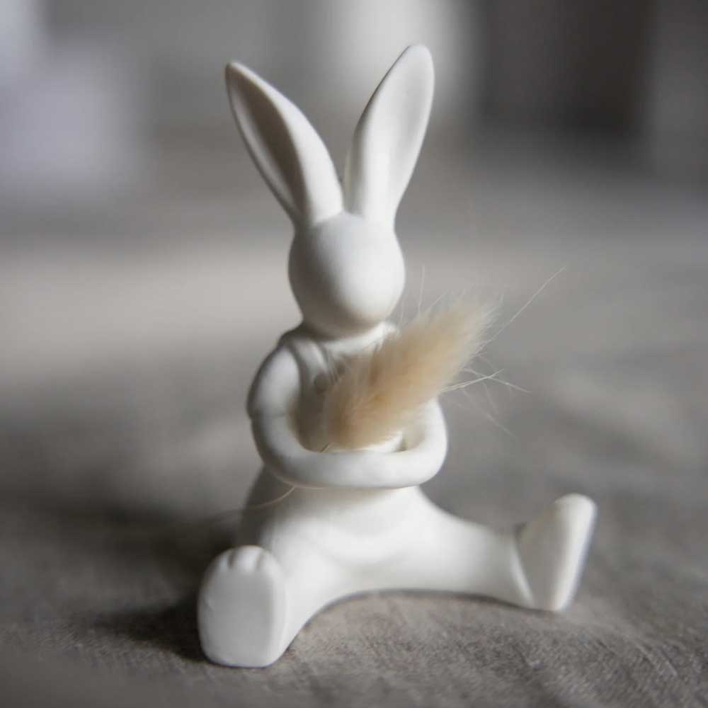 Der Lasse Hase von Storefactory ist eine kleine, weiße Hasenfigur, die sitzend dargestellt ist und einen flauschigen Gegenstand in den Armen hält.