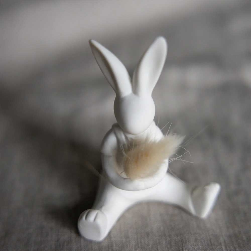 Die „Storefactory – Lasse Hase“ ist eine weiße Hasenfigur aus Keramik mit einem kleinen Büschel beigem Fell auf grauer Stoffoberfläche.