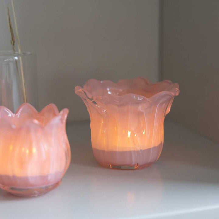 Zwei Storefactory - Ljunga Teelichthalter aus rosa geriffeltem Glas, jeweils mit einer brennenden Kerze darin, stehen auf einem Regal.