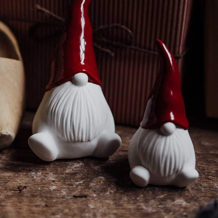 Zwei Weihnachtswichtel aus Keramik von Storefactory, die jeweils eine rote Weihnachtsmütze tragen, stehen auf einer Holzfläche. Ein Wichtel ist größer als der andere. Im Hintergrund sind ein verpacktes Geschenk und ein Teil eines Holzschuhs zu sehen.