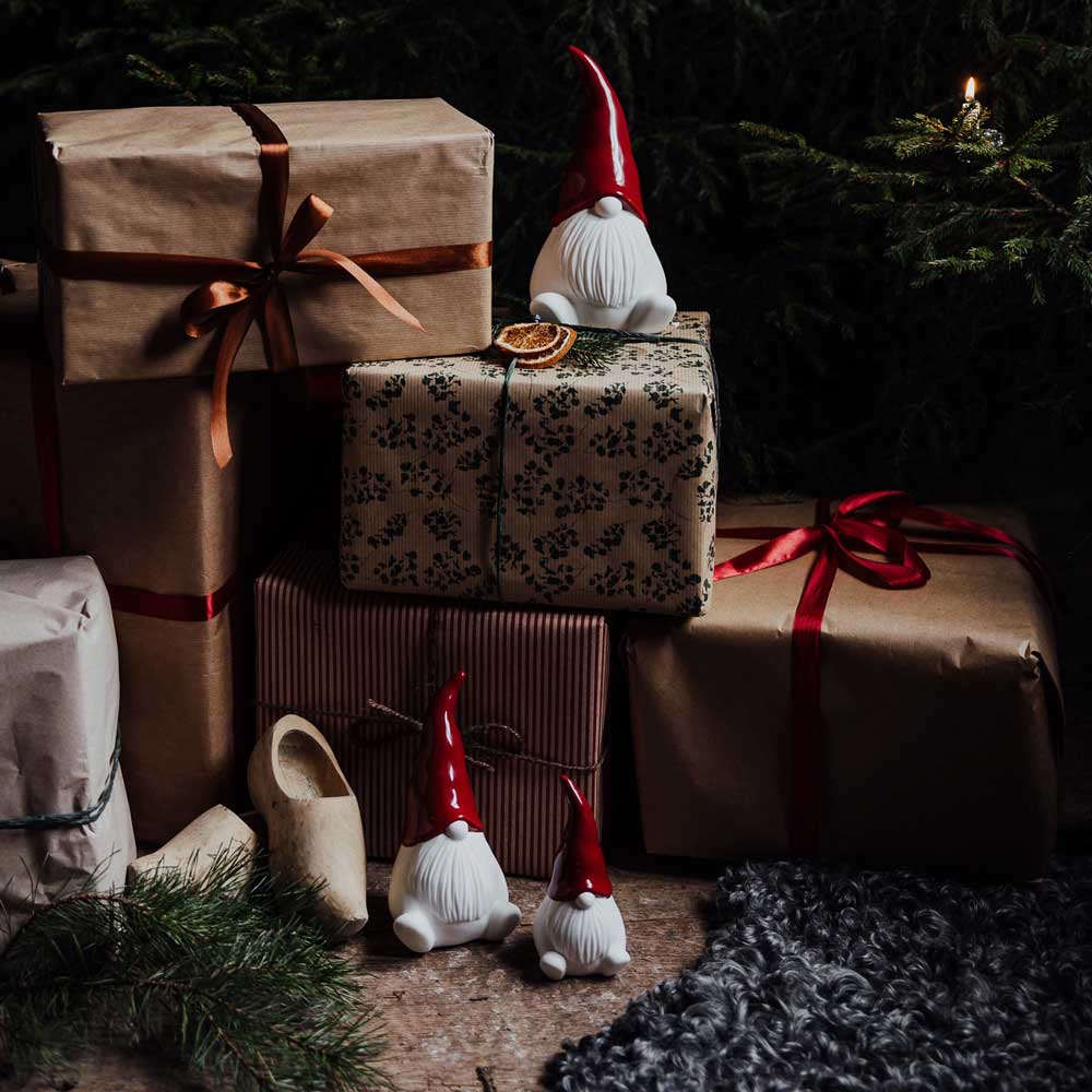 Mit roten und braunen Bändern geschmückte verpackte Geschenke sind auf einem rustikalen Boden gestapelt, begleitet von den Keramikfiguren „Noel Wichtel mit Weihnachtsmütze“ von Storefactory mit roten Hüten neben einem Tannenzweig.