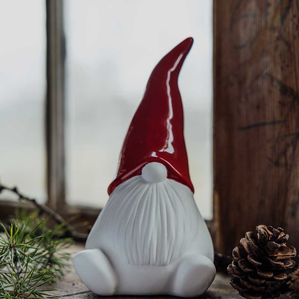 Der Storefactory Noel Wichtel mit Weihnachtsmütze Keramik steht auf einer Holzoberfläche neben einem Tannenzapfen und grünem Laub, mit einem verschwommenen Fenster im Hintergrund.