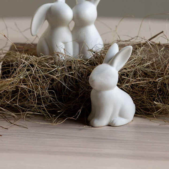 Drei weiße Hasenfiguren aus Keramik von Storefactory, Oskar Hase, sind mit Stroh auf einem hellen Holzuntergrund arrangiert.