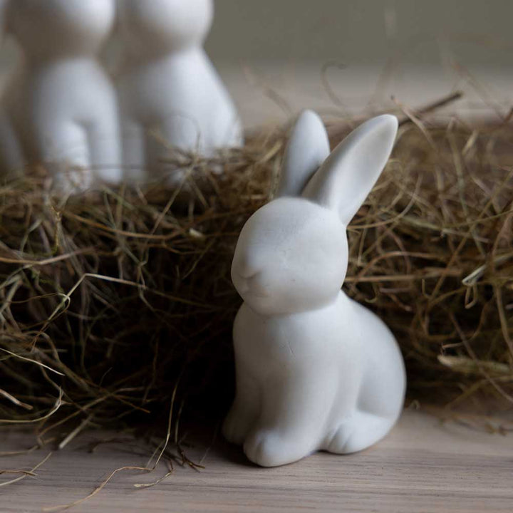 Der Storefactory Oskar Hase, eine kleine weiße Hasenfigur aus Keramik, ruht auf einer Holzoberfläche neben einem Haufen braunem Stroh.