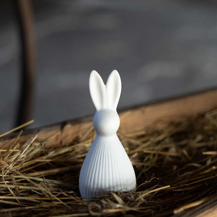 Die Storefactory – Selma Hase ist eine weiße Hasenfigur aus Keramik mit verlängerten Ohren, die auf Stroh in einem Holzbehälter steht.
