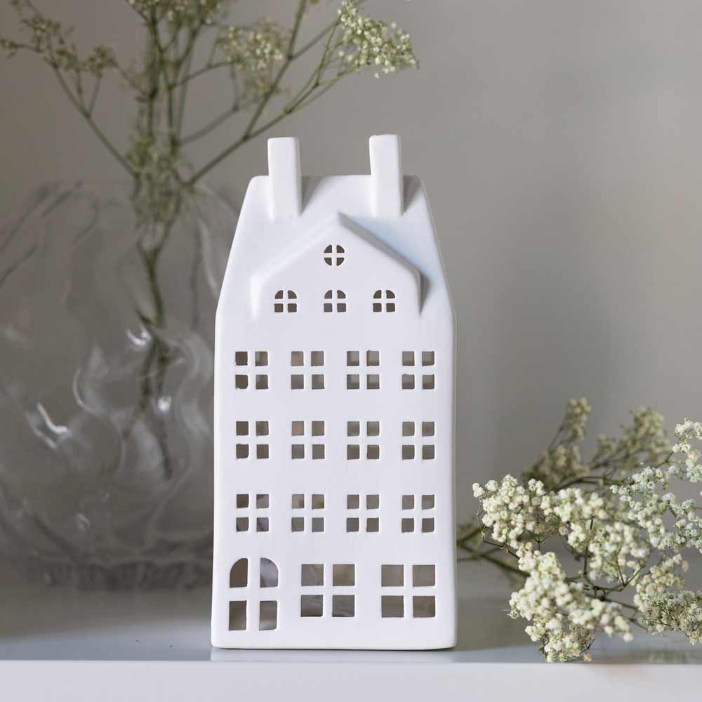 Das Storefactory – Storstaden Nr. 2 Lichthaus Deko, ein weißes Dekohaus aus Keramik, steht auf einer Fläche mit Zweigen zarter weißer Blumen im Hintergrund.