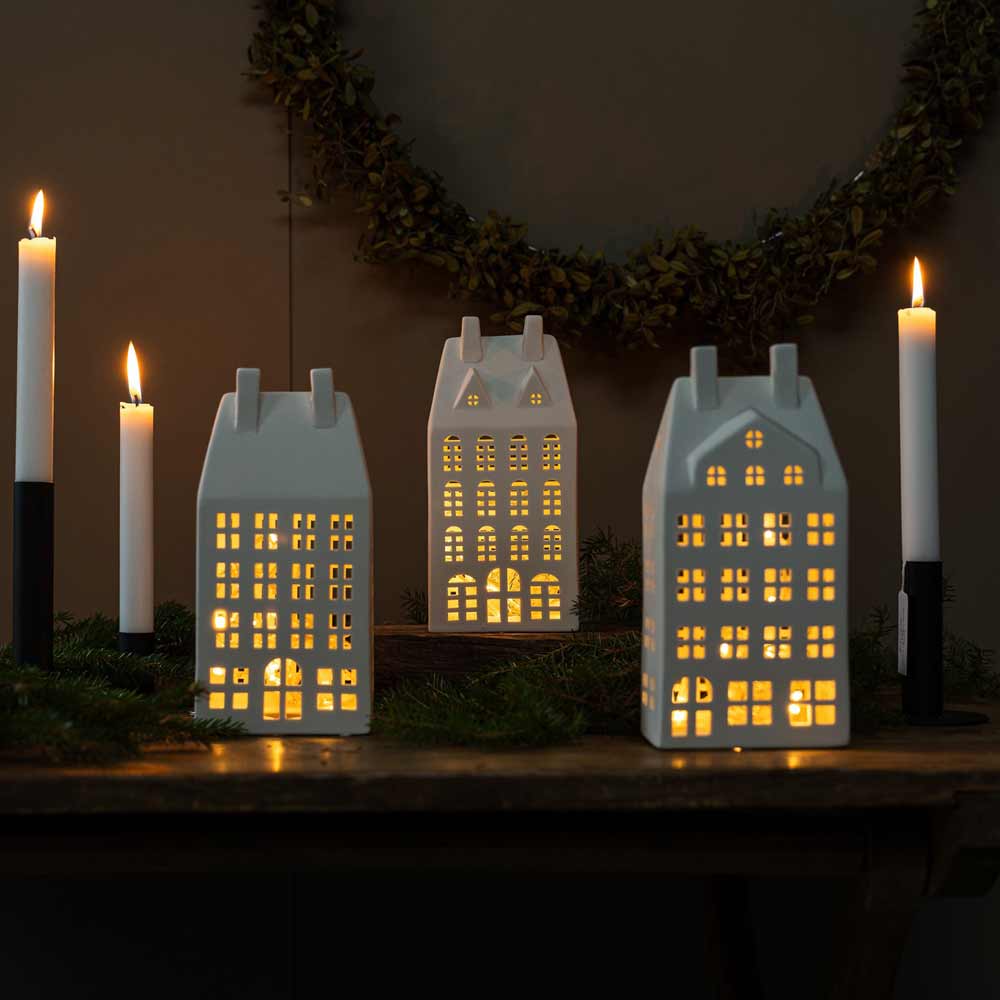 Drei Storefactory – Storstaden Nr. 2 Lichthaus Deko-Laternen und fünf brennende weiße Kerzen sind auf einem Holztisch mit Grün im Hintergrund arrangiert.
