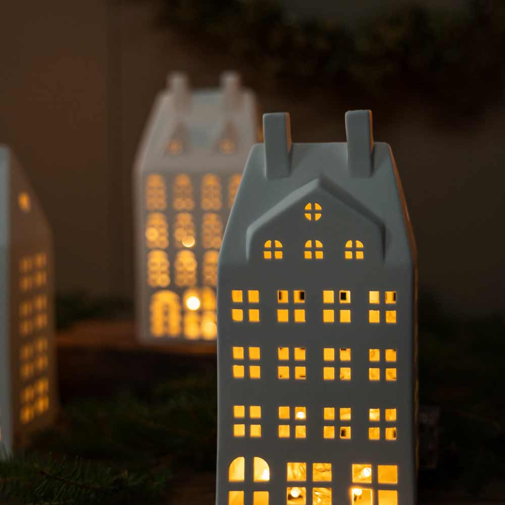 Die Storefactory – Storstaden Nr. 2 Lichthaus Deko, kleine Keramikgebäude mit beleuchteten Fenstern, die an traditionelle europäische Stadthäuser erinnern, sind in einer dunklen Umgebung mit einem verschwommenen Kranz im Hintergrund platziert.