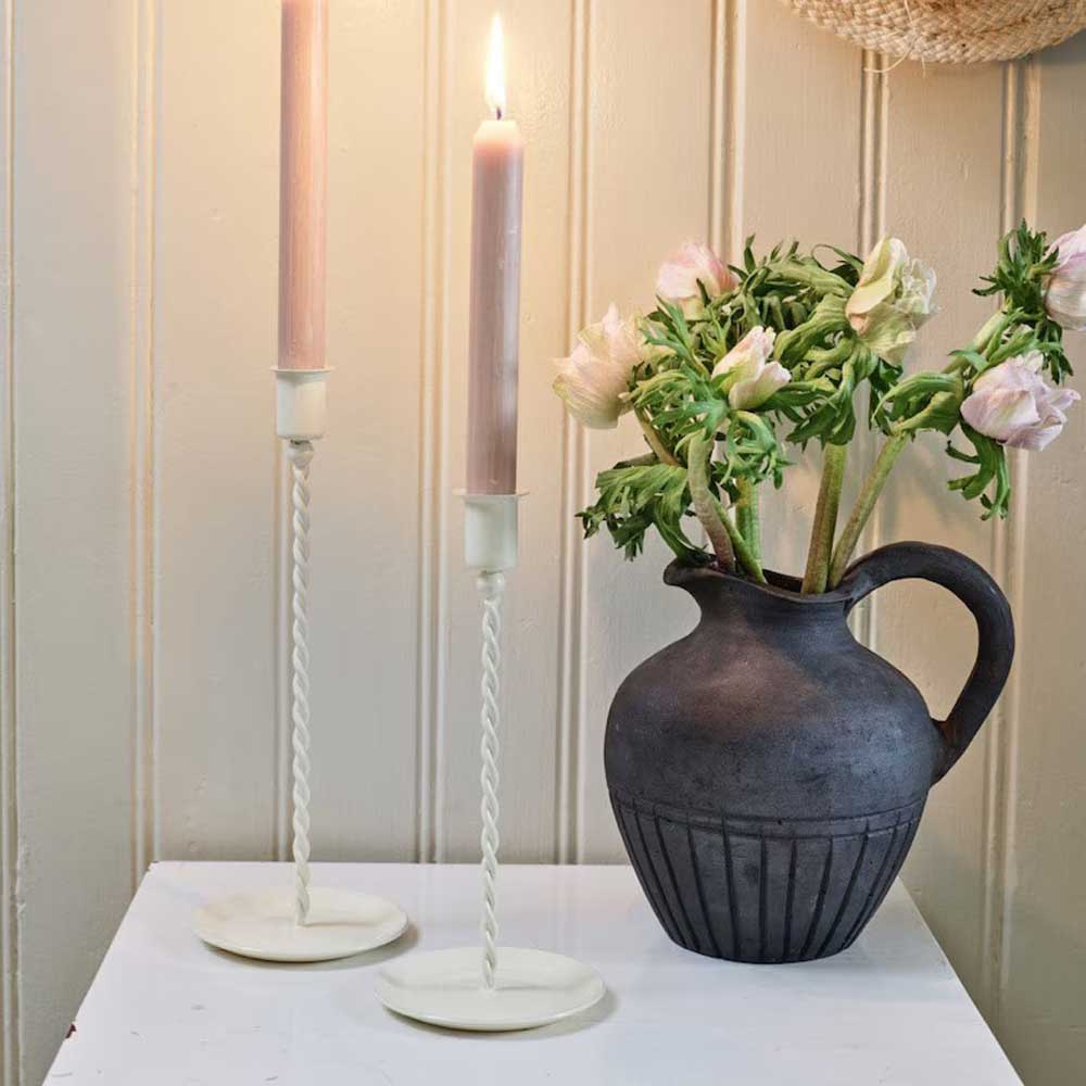 Zwei schlanke weiße Kerzenhalter mit rosa Kerzen neben einem dunklen bauchigen Blumenkrug auf einer weißen Ablage