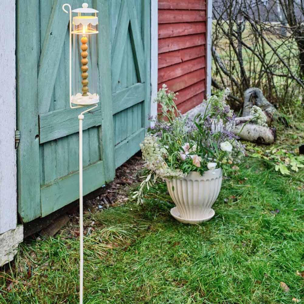 Eine Strömshaga - Laterne Inga auf Gartenspiess steht neben einer grünen Tür einer Holzkonstruktion. Auf dem Rasen daneben steht ein großer Blumentopf mit verschiedenen Blumen.
