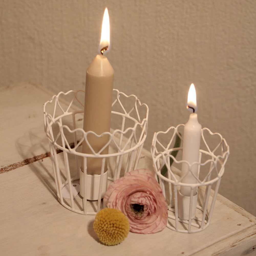 Das VL Home - Kerzenhalter Töpfchen Herz 2er-Set besteht aus zwei brennenden Kerzen in weißen Drahthaltern mit Herzmotiven, die auf einer weißen Oberfläche neben einer rosa Ranunkel und einer gelben Billyball-Blume präsentiert werden.