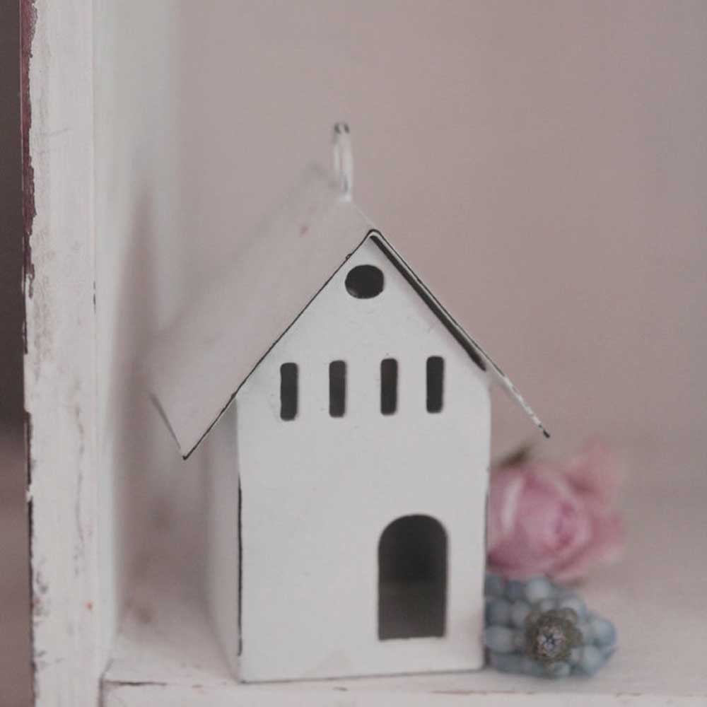 Auf einem Regal steht ein kleines, weißes, metallisches Haus-Ornament mit Spitzdach und schwarzen ausgeschnittenen Fenstern. Neben dem VL Home - Metallhaus Deko weiß Mini unten geöffnet liegen eine rosa Rose und hellblaue Perlen.