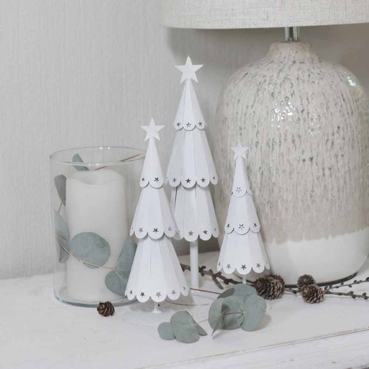 Der VL Home – Tannenbaum Metall mit Sternen und ein mit Eukalyptusblättern geschmückter Kerzenhalter aus Glas sind auf einer weißen Oberfläche neben einer strukturierten Lampe arrangiert.