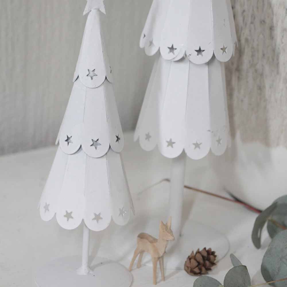 Zwei weiße Bäume mit Sternenmuster von VL Home – Tannenbaum Metall mit Sternen, begleitet von einem kleinen hölzernen Hirsch und einem Tannenzapfen auf einem Tisch.