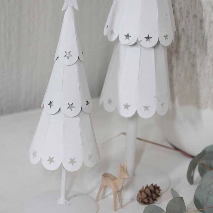 Zwei weiße Bäume mit Sternenmuster von VL Home – Tannenbaum Metall mit Sternen, begleitet von einem kleinen hölzernen Hirsch und einem Tannenzapfen auf einem Tisch.