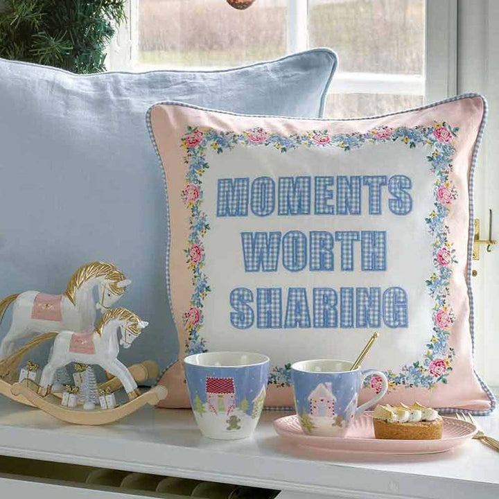 Dekoratives Kissen mit der Stickerei „Moments Worth Sharing“, daneben ein hölzernes Schaukelpferd und zwei Tassen Tee mit Scones auf einem Tablett und die GreenGate - Alice Servierplatte.