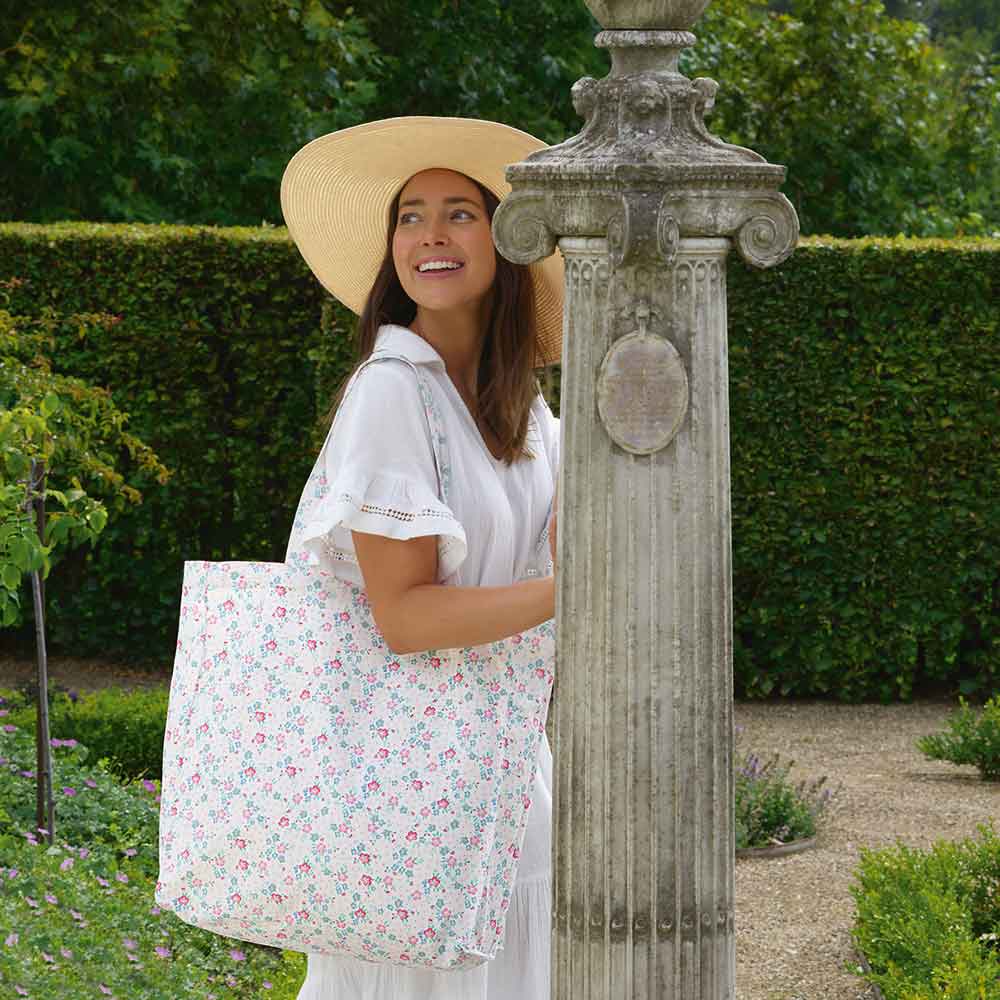 Eine Frau in einem weißen Kleid und einem breitkrempigen Hut steht neben einer Steinsäule in einem Garten und hält die GreenGate - Allyna Geschirrtuch White Einkaufstasche.