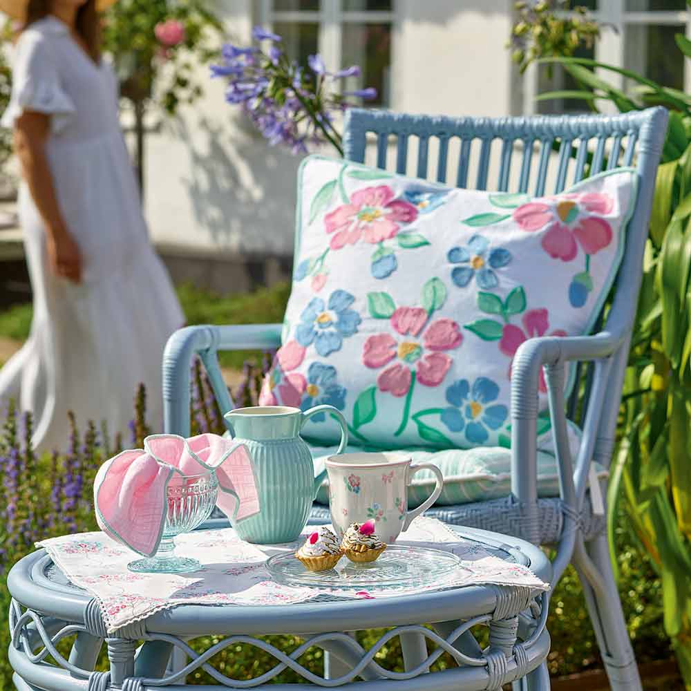 Ein blauer Stuhl mit einem geblümten Kissen steht neben einem runden Tisch, auf dem ein Krug, eine Tasse, ein Kuchen und eine weiße Serviette von Allyna Geschirrtuch stehen. Im Hintergrund geht eine Person in einem weißen Kleid durch den Garten.