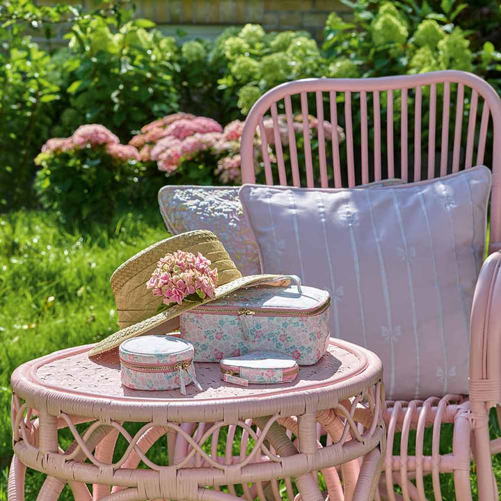 Ein rosa Korbstuhl und ein Tisch in einem Garten mit einem Strohhut mit einer Blume, geblümten Stoffboxen und einem Kissen. Das GreenGate - Allyna Geschirrtuch in Weiß verleiht dem Tisch, umgeben von üppigem Grün mit Sträuchern und blühenden Blumen, Eleganz.