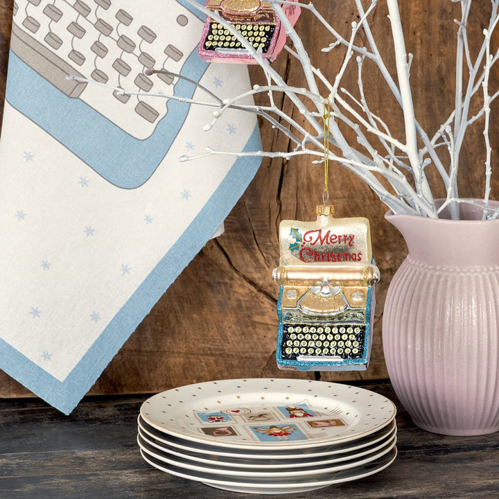 Ein GreenGate Xmas Letter Sticker-Set White schmückt eine weiße Vase mit Zweigen, die neben dekorierten Tellern und einem Handtuch mit Schreibmaschinendesign auf einem Holztisch steht.