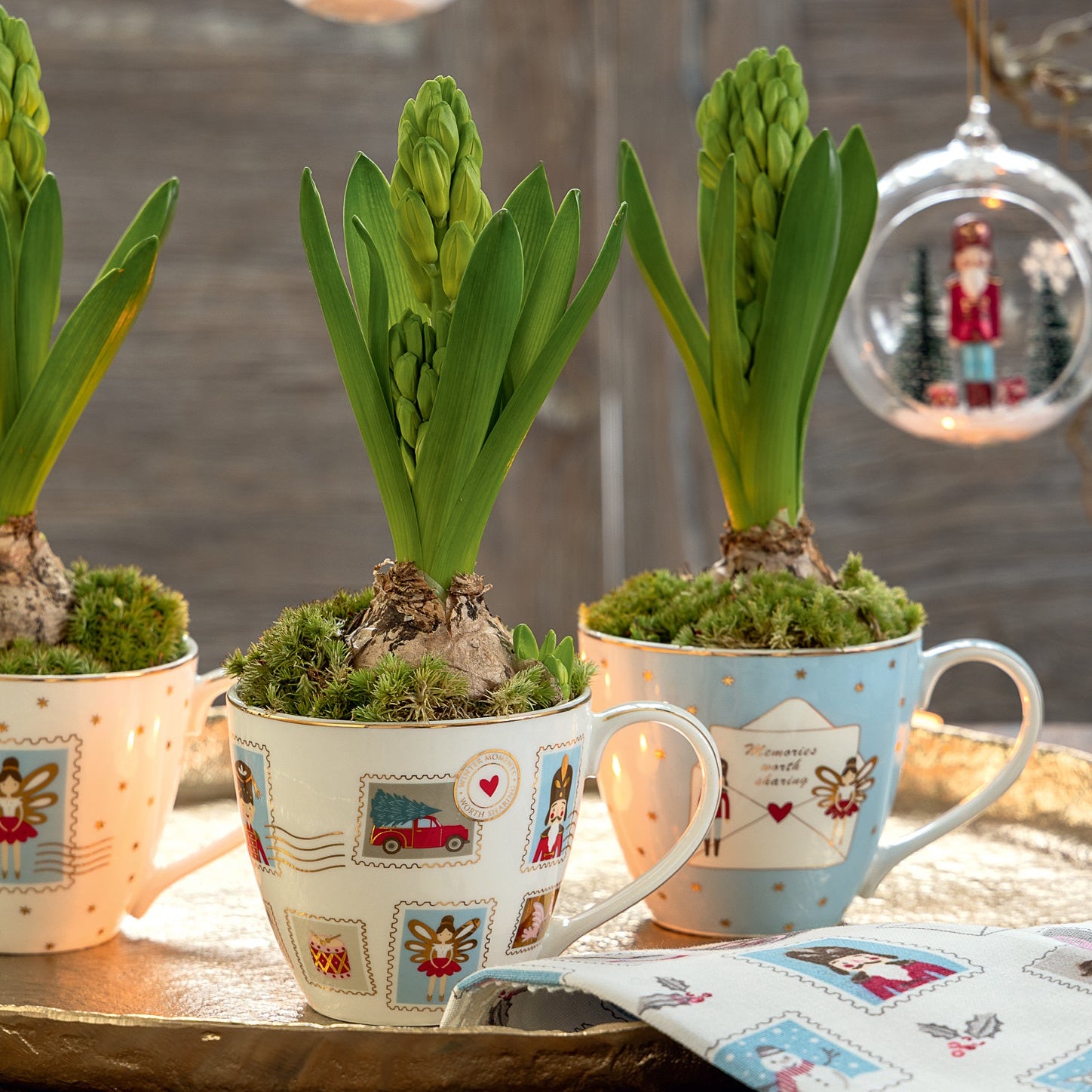 Drei Teetassen mit festlichen Motiven halten grüne Hyazinthen in Töpfen auf einem Tablett, begleitet vom GreenGate - Xmas Letter Kalender quadratisch White und einer Serviette.