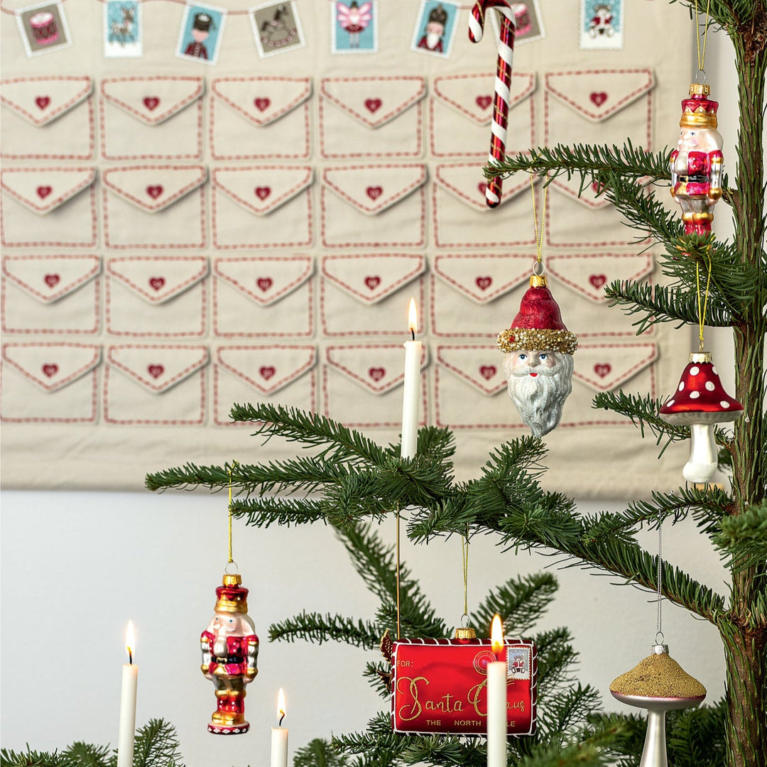 Ein geschmückter Weihnachtsbaum mit Kerzen, Nussknacker-Ornamenten, Weihnachtsmannkopf und einem roten Briefkasten-Ornament. Im Hintergrund hängt der GreenGate - Xmas Letter Kalender quadratisch weiß mit Umschlagtaschen für die Adventszeit.