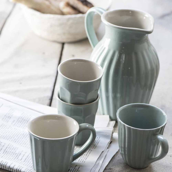 Eine Sammlung von Ib Laursen - Cafe Latte Becher Mynte-Tassen und ein Krug in Pastellgrün auf einem Holztisch, mit einer Zeitung und einem Brotkorb im Hintergrund.