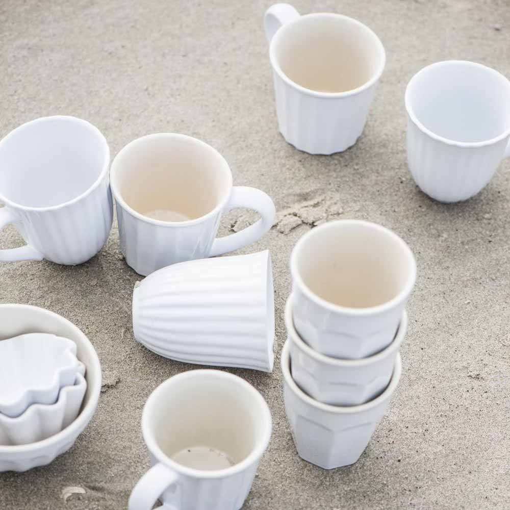 Mehrere weiße Ib Laursen - Cafe Latte Becher Mynte Becher und Schüsseln, teilweise gestapelt, auf einem sandigen Untergrund.