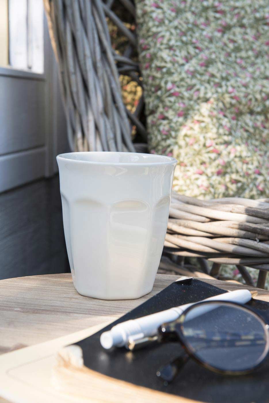 Ib Laursen - Cafe Latte Becher Mynte auf einem Holztisch mit Sonnenbrille und einem Notizbuch daneben, Außenambiente mit Korbgeflecht und floralem Hintergrund.