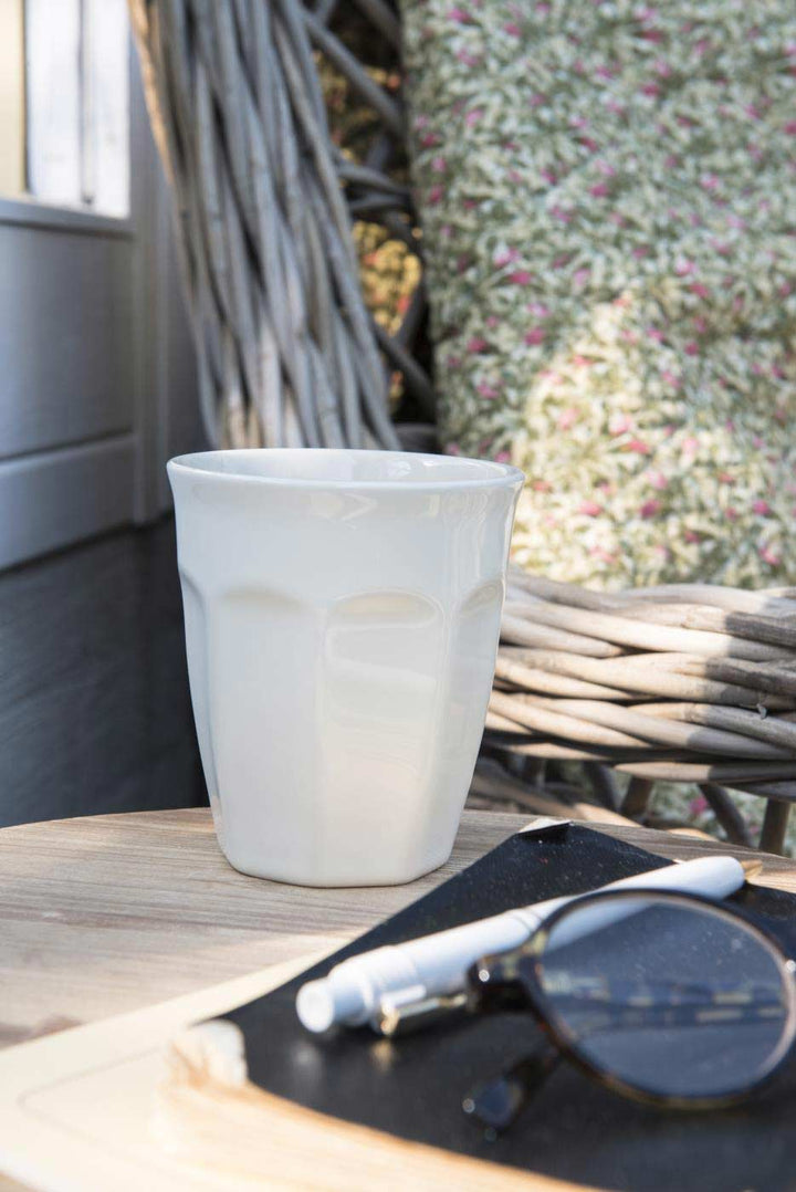 Ib Laursen - Cafe Latte Becher Mynte auf einem Holztisch mit Sonnenbrille und einem Notizbuch daneben, Außenambiente mit Korbgeflecht und floralem Hintergrund.