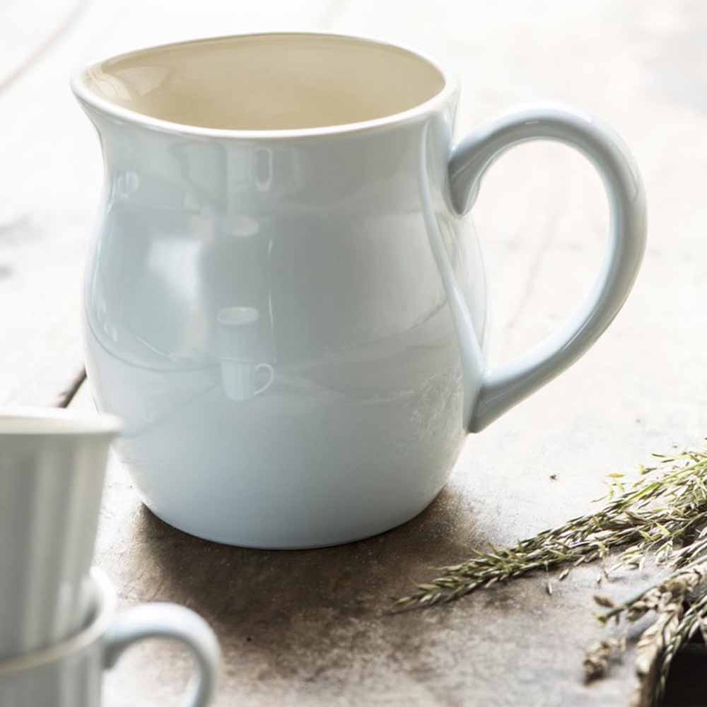 Eine schlichte, hellblaue Ib Laursen - Kanne 2,5 Liter Mynte auf einer Betonfläche, teilweise gefüllt mit einer beigen Flüssigkeit, neben einem kleinen Bündel getrockneter Kräuter.