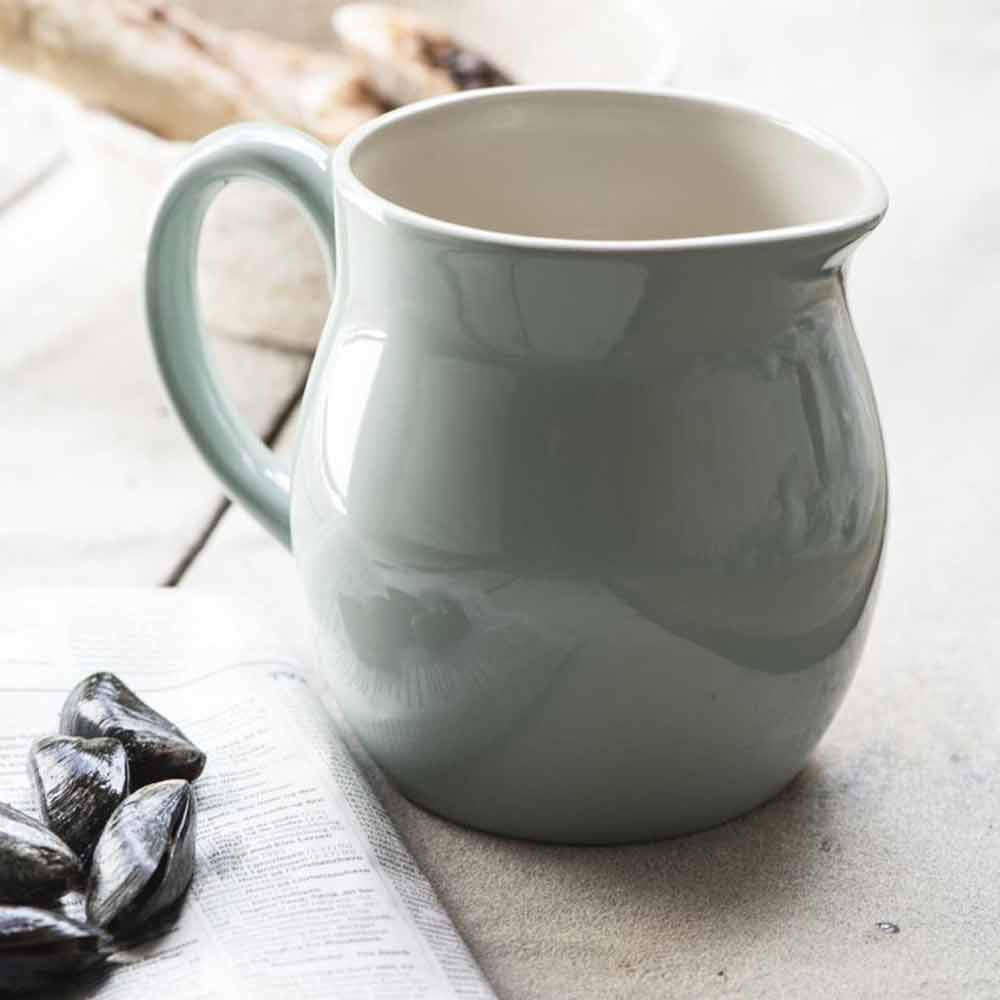 Eine Ib Laursen - Kanne 2,5 Liter Mynte Keramiktasse auf einem Tisch neben Sonnenblumenkernen und einem teilweise sichtbaren offenen Buch.