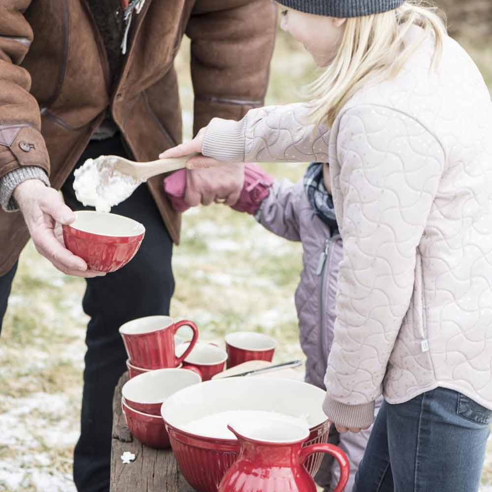 Eine Person serviert einer anderen Person Essen im Freien. Dabei verwendet sie einen Holzlöffel und Ib Laursen - Krug Mynte 1,7-Liter-Schüsseln. Weitere Gerichte stehen auf einem rustikalen Tisch.