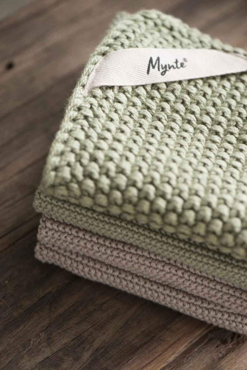 Ein Stapel ordentlich gefalteter grün-beiger Strickhandtücher mit einem sichtbaren Etikett mit der Aufschrift „Ib Laursen – Topflappen Mynte“ auf einer Holzoberfläche.