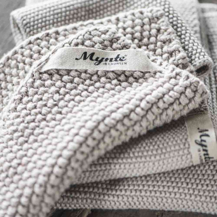 Nahaufnahme eines Stapels grauer Stricktextilien mit einem sichtbaren Etikett mit der Aufschrift „Ib Laursen – Topflappen Mynte“.