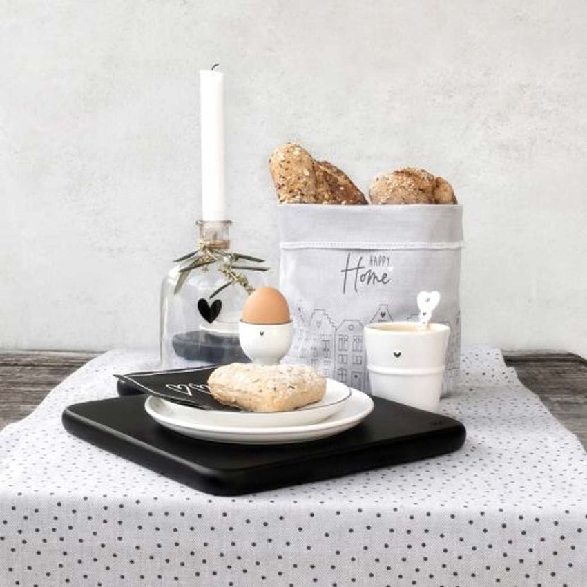Frühstücksanordnung mit Brötchen, Ei, weißem Herzbecher und Brotkorb auf einem schwarz-weiß gedeckten Tisch