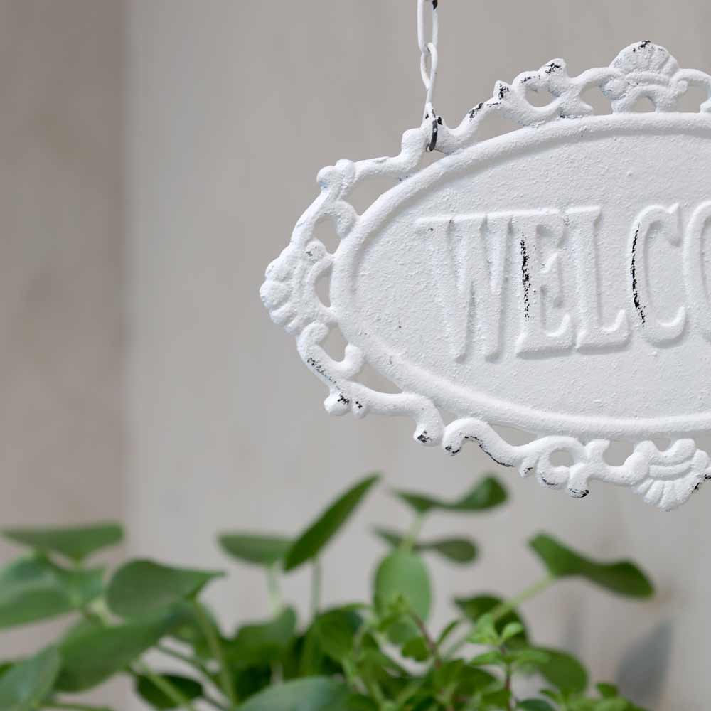 Chic Antique - Schild „Willkommen“ hängt an einer Wand, im Hintergrund sind sanft verschwimmende grüne Pflanzenblätter zu sehen.