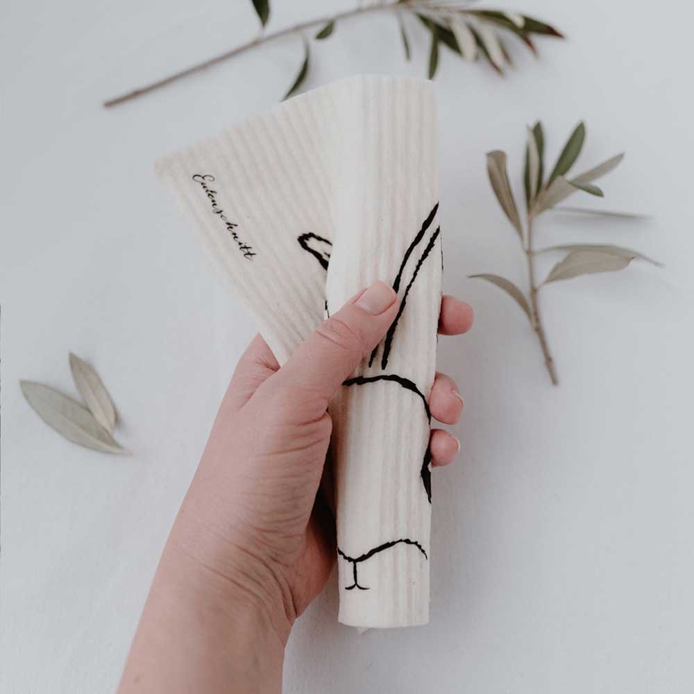 Eine Hand hält drei gefaltete Eulenschnitt - Schwammtücher Hasi 3er Set mit minimalistischem Vogeldesign, vor einem neutralen Hintergrund mit verstreuten Blättern.