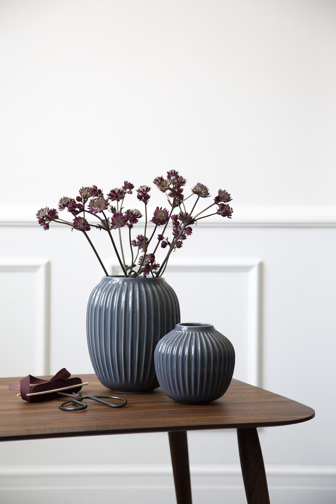 Zwei anthrazitfarbene gerillte Vasen auf einem Holztisch vor einer hellen Wand; eine Vase trägt dunkelrote Blüten