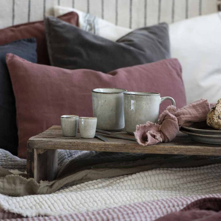 Zwei große Keramikbecher und zwei Ib Laursen - Eierbecher Sand Dunes auf einem Holztablett mit einem Laib Brot, auf einem Bett mit einer gestreiften Decke und Kissen.